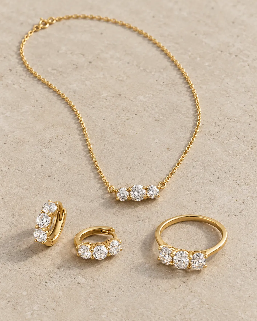 Gold three-stone diamond necklace, huggie earrings and ring on a stone surface